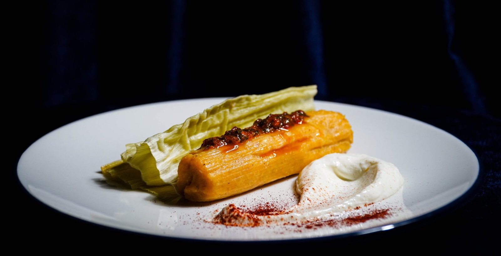 Tamal servido en plato blanco sobre terciopelo azul oscuro, con salsa y crema con pimentón